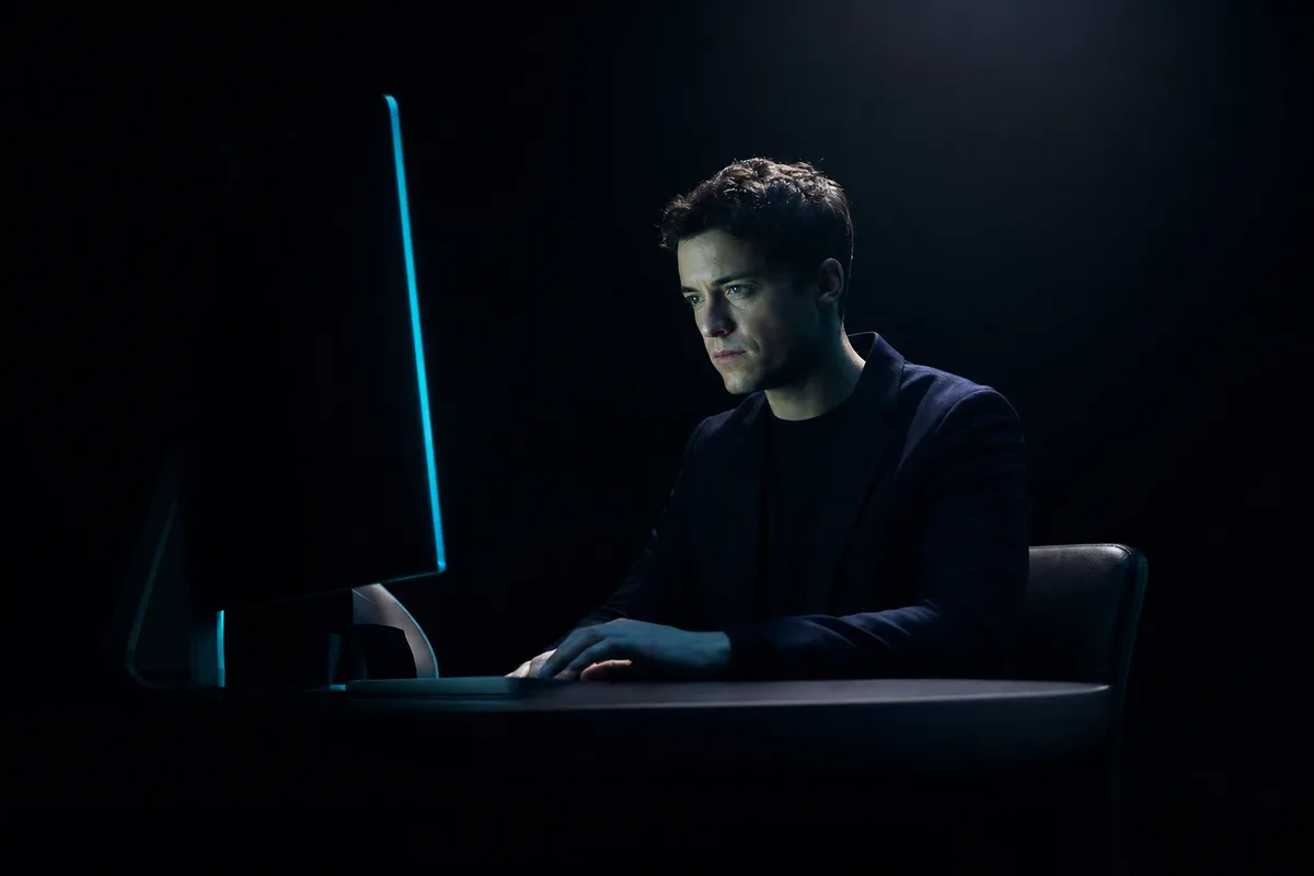 A professional at a dark minimal desk, face lit by a single electric cyan monitor glow — the experience of working alongside a proactive AI agent